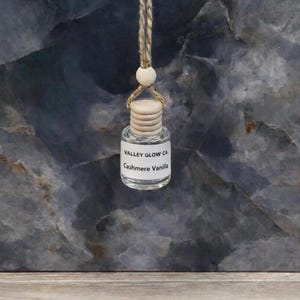 May include: A small, clear glass bottle with a wooden cap and a beige string for hanging. The bottle has a white label with the text "VALLEY GLOW CA" and "Cashmere Vanilla". The background is a dark blue and gray marbled surface.