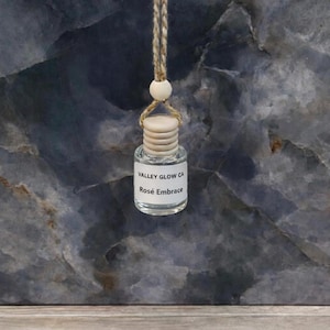 May include: A small, clear glass bottle with a wooden cap and a label that reads "Valley Glow CA Rosé Embrace." The bottle is suspended by a natural fiber string with a wooden bead. The background is a dark, textured surface.