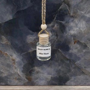 May include: A small, clear glass bottle with a wooden cap and a string for hanging. The label reads "VALLEY GLOW CA" and "After Hours". The background is a dark, marbled surface.