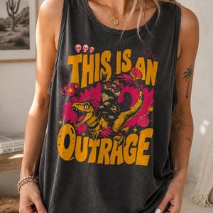 May include: A black sleeveless tank top with a graphic design featuring a dinosaur and the words "THIS IS AN OUTRAGE" in yellow. The design has pink and yellow accents. The tank top is made of a soft, comfortable material.