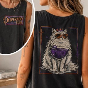 May include: Dark gray tank top featuring a graphic of a fluffy cat wearing sunglasses and holding a purple cocktail with a cherry. The tank top also has the text "The Desperado Fun It Hurts Club" on the front.