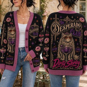 K&ouml;nnte beinhalten: Eine schwarze Strickjacke mit violetten R&auml;ndern und einer Grafik einer Katze mit Sonnenbrille neben einem Cocktailglas. Auf der R&uuml;ckseite steht der Text "The Desperado Club" und "Dirty Shirley" mit Abbildungen von Schwertern, Kirschen und Donuts.