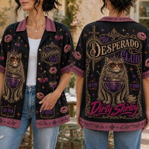 K&ouml;nnte beinhalten: Ein schwarzes kurz&auml;rmeliges Hemd mit Knopfleiste und einer Grafik einer Katze mit Sonnenbrille und Krone, die einen lila Cocktail h&auml;lt. Das Design enth&auml;lt den Text The Desperado Club, So Fun, It Hurts und Dirty Shirley sowie Kirsch- und Donut-Motive.