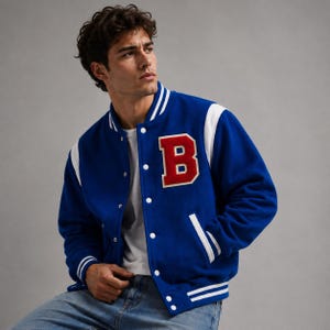 May include: A royal blue varsity jacket with white accents on the shoulders, collar, and cuffs. The jacket features a large red "B" patch on the chest and white snap closures. The jacket is worn by a person.