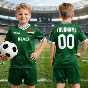 K&ouml;nnte beinhalten: Ein Kind in einem gr&uuml;nen Fu&szlig;balltrikot mit wei&szlig;en Details, das einen Fu&szlig;ball h&auml;lt. Das Trikot zeigt die irakische Flagge, das Jahr 2026 und das Wort "IRAQ". Auf der R&uuml;ckseite des Trikots stehen "YOURNAME" und die Nummer "00".