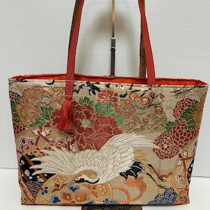 以下が含まれることがあります： 鶴と花の模様が施されたベージュのトートバッグ。赤いハンドルと赤いタッセルが特徴です。デザインには金、赤、緑、オレンジの色が使われており、細部までこだわった華やかな印象です。内側はオレンジ色の生地で裏打ちされています。