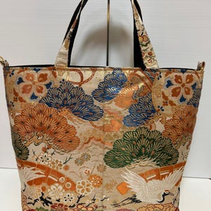 以下が含まれることがあります： オレンジ、青、緑の花柄と鶴柄が施されたベージュのトートバッグ。バッグは黒いハンドルと黒い内側を備えています。デザインは、様式化された木、花、鳥を特徴としています。