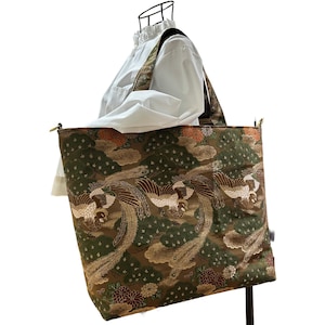 May include: A large tote bag with a detailed pattern featuring phoenixes, flowers, and clouds in shades of green, brown, and gold. The bag has long straps and a rectangular shape, suitable for carrying various items.