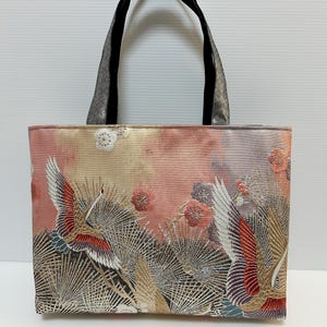 以下が含まれることがあります： 鶴と花の要素をあしらった装飾的なデザインの長方形トートバッグ。バッグは、ピーチ、ゴールド、グレーの色使いで、黒いハンドルが付いています。鶴は赤、白、金のディテールが施されています。バッグはテクスチャード加工された生地で作られています。