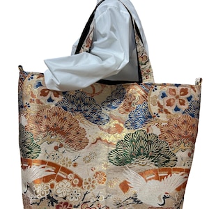 May include: Large tote bag with a cream-colored background and a detailed pattern featuring cranes, flowers, and pine trees in shades of orange, blue, and green. The bag has black straps and a white interior.