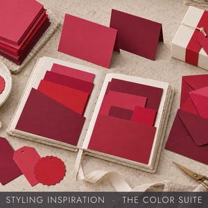 May include: A collection of red stationery items including a stack of cards, a small open book with pockets holding various red paper swatches, gift tags, envelopes, and a gift box wrapped with a red ribbon and a tag. Gold scissors are included.