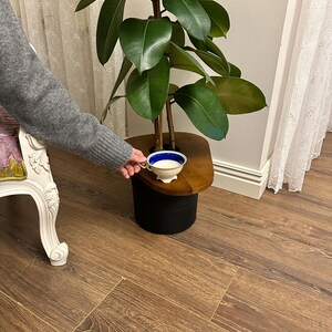 K&ouml;nnte beinhalten: Eine kleine, wei&szlig;e Teetasse mit blauem Rand steht auf einem h&ouml;lzernen Beistelltisch neben einer gro&szlig;en Pflanze mit gr&uuml;nen Bl&auml;ttern. Der Tisch wird von einem schwarzen Sockel getragen. Der Boden ist ein warmer, brauner Laminatboden in Holzoptik.