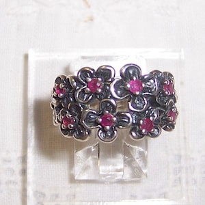May include: A silver ring with a floral design featuring multiple pink gemstones. The ring is displayed on a clear plastic stand.