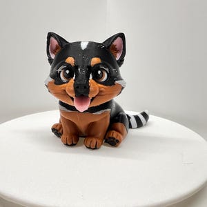 May include: A 3D-printed figurine of a puppy, featuring a black and tan coat with a white spot on its head. The puppy is sitting with its tongue sticking out, showcasing a playful expression. The figurine is on a white, round surface.