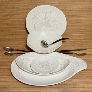May include: A cream-colored dinnerware set with a floral design. The set includes plates, bowls, and serving pieces. The floral design features gray lines and pink dots. Silverware with wooden handles is also included.