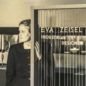 May include: Sepia-toned photograph featuring a woman standing near a screen with vertical lines. The screen displays the text "EVA ZEISEL INDUSTRIAL DESIGNER" in white block letters. The woman is wearing a dark-colored dress.