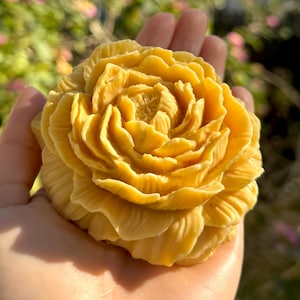 Puede incluir: Una flor tallada amarilla detallada, posiblemente jabón o cera, sostenida en una mano. La flor tiene muchas capas de pétalos, con una superficie texturizada. El fondo está borroso, con follaje verde y rosa.