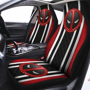 May include: Car seat covers featuring a black and red design with white stripes. The covers have a Deadpool logo in the center and on the headrests. The seats are in a car interior.