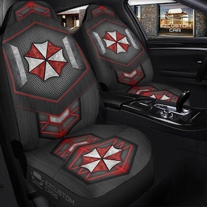 May include: Car seat covers with a black and gray design featuring a red and white umbrella logo. The covers have a geometric pattern with red accents and are visible inside a car.