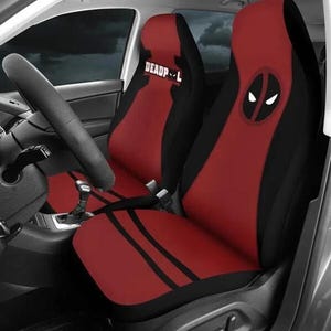 May include: Red and black car seat covers featuring the Deadpool logo and text. The seat covers have a red center with black accents and stripes. The Deadpool logo is a white eye design on a black circle.