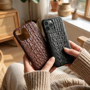 May include: Two smartphone cases with a textured crocodile skin pattern. One case is dark brown and the other is black. Both cases feature a rectangular cutout for the camera lens array on the back, showcasing a detailed embossed surface.