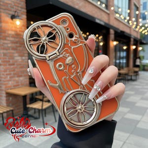 May include: Orange phone case with a silver-colored decorative design and two circular, wheel-like features. The case has a built-in stand. The words "Cute Charm corner" are in the bottom left corner.