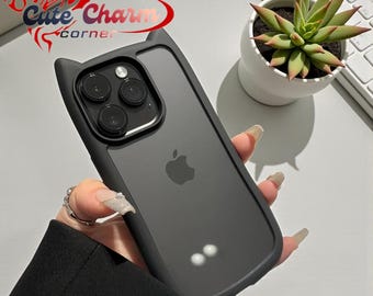 May include: A black iPhone case with cat ear design. The case has a clear back and a black frame. The case is held in a hand. The image also shows a succulent plant in a white pot and a computer keyboard.