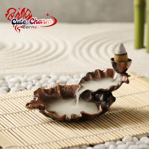 Puede incluir: Un quemador de incienso de color cobre en forma de hoja, con un pequeño palito de incienso cónico encendido. El quemador emite humo blanco, creando un ambiente relajante. El texto "Cute Charm Corner" es visible.