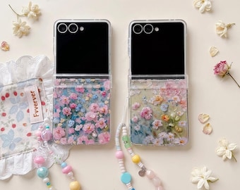 May include: Two clear phone cases with floral designs, featuring pink and blue flowers. Each case has a black screen and a beaded strap with colorful beads. The cases are on a light background with scattered flowers.