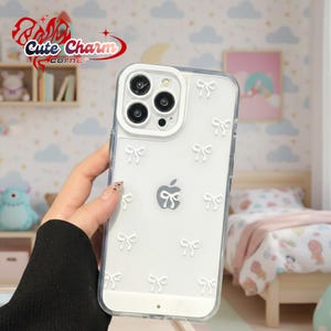 Puede incluir: Funda transparente para teléfono con lazos blancos. La funda presenta un diseño transparente con un patrón de lazos blancos y un logotipo de Apple. El texto "Cute Charm Corner" es visible en la esquina superior izquierda.