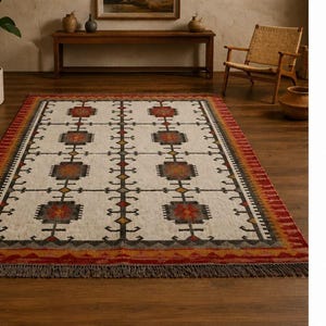 Puede incluir: Una alfombra rectangular con base color crema y un patrón geométrico repetitivo en tonos gris carbón, naranja óxido y amarillo mostaza. La alfombra tiene un borde ancho en tonos rojos y naranjas y está rematada con flecos gris oscuro.