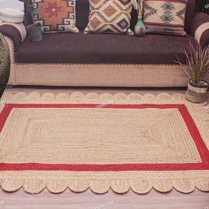 May include: A rectangular area rug crafted from natural tan jute fibres, featuring a bold red rectangular border and a decorative scalloped edge. The rug has a woven texture and is laid flat, showcasing a consistent pattern throughout its surface.
