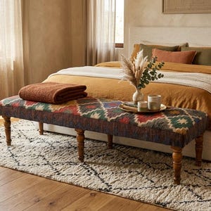 May include: A bedroom scene with a bed featuring mustard-coloured bedding and decorative cushions. A patterned bench with wooden legs is at the bed's foot. A folded brown blanket rests on the bench. A rug and hardwood floor complete the scene.