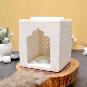 Può includere: Un portacandele in marmo bianco con un ritaglio ad arco e un intricato design a griglia. Una piccola candela accesa si trova all'interno del portacandele a forma di cubo. Il portacandele è su una base di legno.