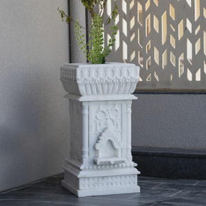 May include: A white marble planter with intricate carvings and a square top. The planter features detailed floral and geometric patterns, and is filled with green plants. The design is ornate and elegant, suitable for indoor or outdoor use.