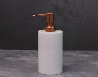 May include: A white marble soap dispenser with a copper-coloured pump. The cylindrical dispenser has a smooth, polished surface. The pump has a curved spout. The dispenser is designed for use in a bathroom or kitchen.