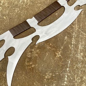 May include: A large, decorative metal blade with a polished silver finish. The blade features a series of curved cutouts and is accented with brown leather-wrapped sections. The design suggests a fantasy or medieval theme.