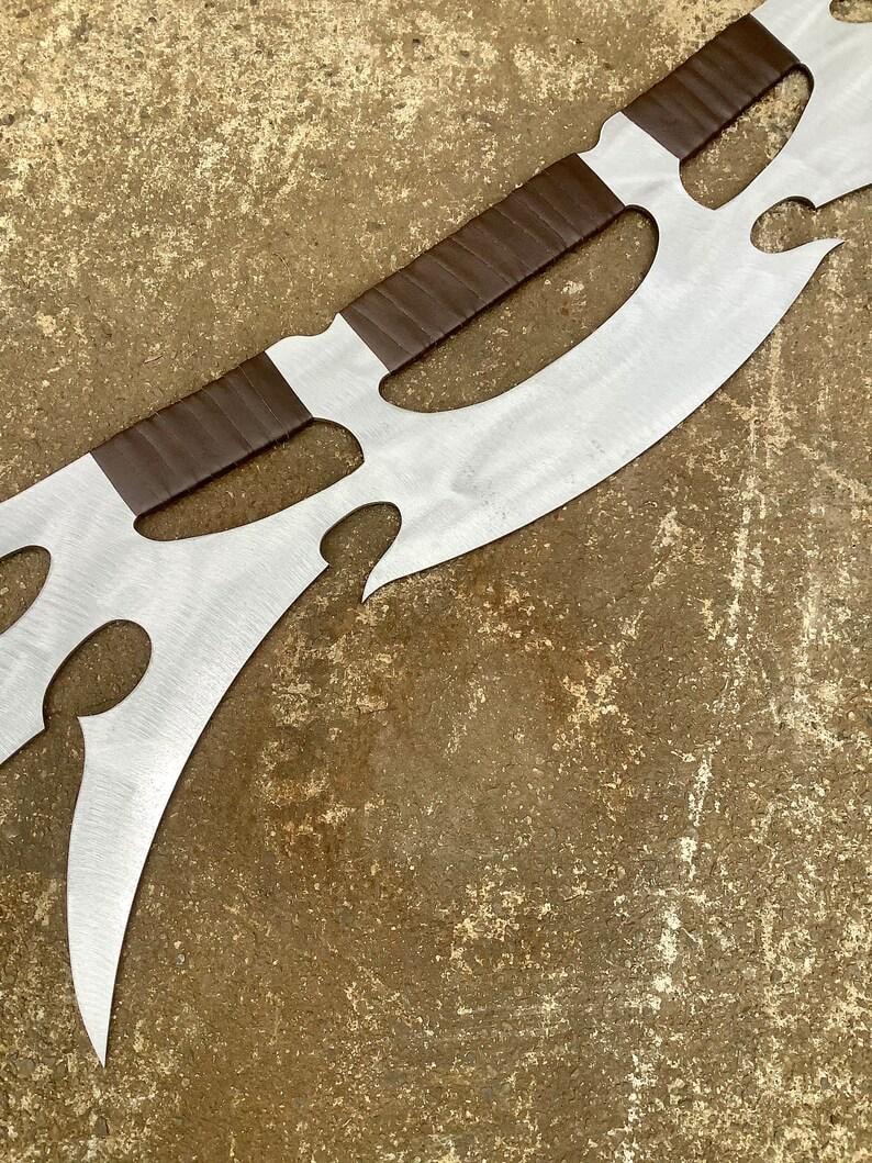 May include: A large, decorative metal blade with a polished silver finish. The blade features a series of curved cutouts and is accented with brown leather-wrapped sections. The design suggests a fantasy or medieval theme.