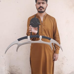 May include: A prop replica of a Klingon Bat'leth, a curved, double-bladed sword. The weapon features a silver blade with black handles and a black emblem. The prop is held in front of a person wearing a brown outfit.