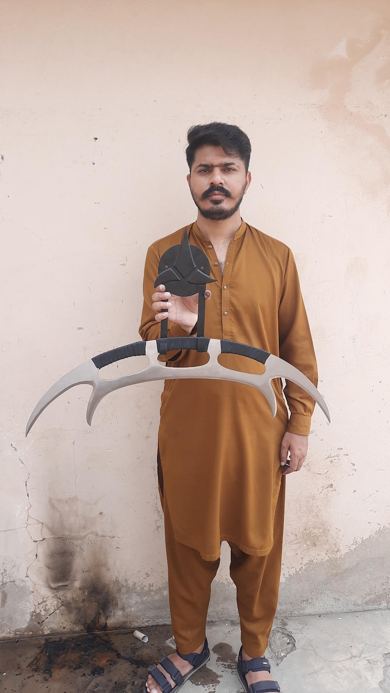 May include: A prop replica of a Klingon Bat'leth, a curved, double-bladed sword. The weapon features a silver blade with black handles and a black emblem. The prop is held in front of a person wearing a brown outfit.