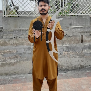 May include: A person holding a black hat and a silver and black decorative blade. The blade has a curved design with black accents. The person is wearing a mustard-colored outfit and black sandals.