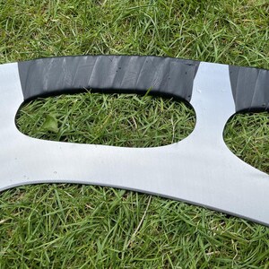 May include: A silver-colored, curved metal object with a black grip. The object has a unique shape with two large, open areas. The item is resting on a bed of green grass.