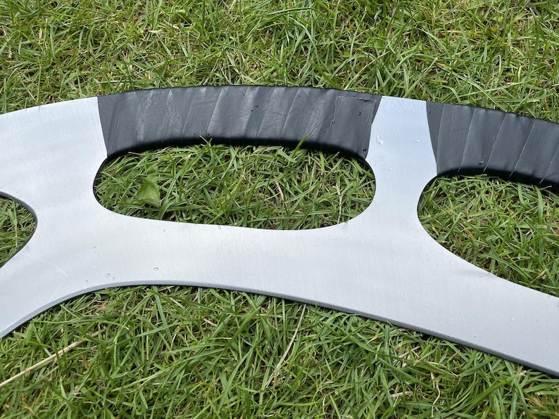 May include: A silver-colored, curved metal object with a black grip. The object has a unique shape with two large, open areas. The item is resting on a bed of green grass.