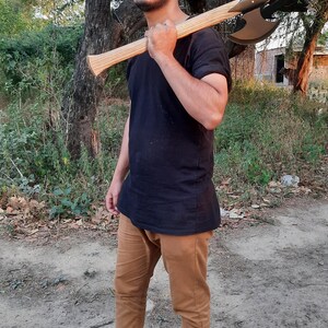Puede incluir: Un hacha de batalla de doble filo con una cabeza de color gris met&aacute;lico y un mango de madera clara, apoyada sobre un hombro. El hacha presenta un dise&ntilde;o de hoja curva y sim&eacute;trica con un punto de montaje central oscuro que une la cabeza al mango.