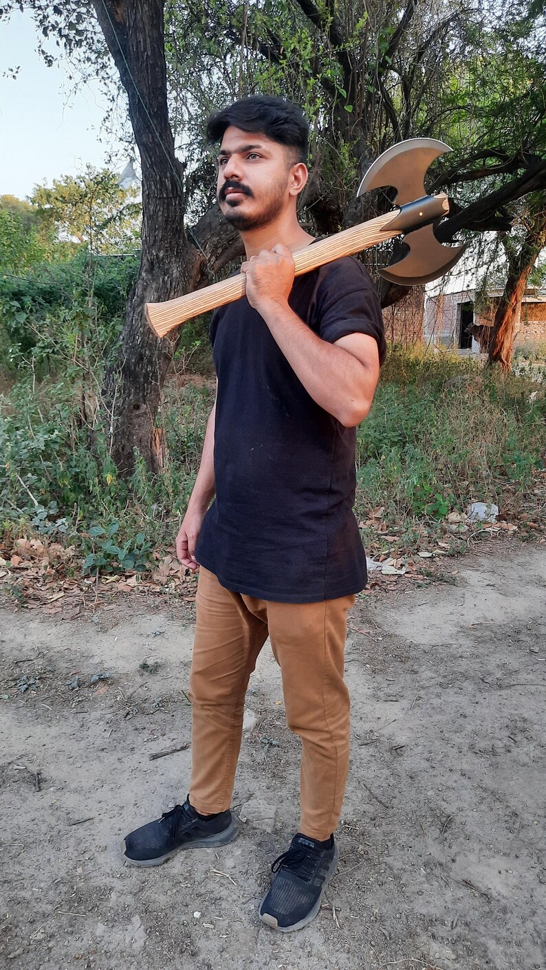 Puede incluir: Un hacha de batalla de doble filo con una cabeza de color gris met&aacute;lico y un mango de madera clara, apoyada sobre un hombro. El hacha presenta un dise&ntilde;o de hoja curva y sim&eacute;trica con un punto de montaje central oscuro que une la cabeza al mango.