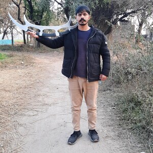 May include: A person holding a large, silver-colored, curved blade weapon. The weapon has a unique design with multiple curved edges. The person is wearing a black jacket, a blue shirt, and tan pants. The background shows trees and a dirt path.