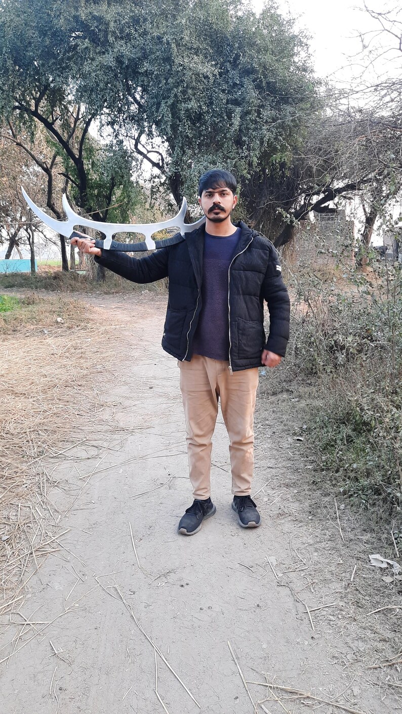 May include: A person holding a large, silver-colored, curved blade weapon. The weapon has a unique design with multiple curved edges. The person is wearing a black jacket, a blue shirt, and tan pants. The background shows trees and a dirt path.