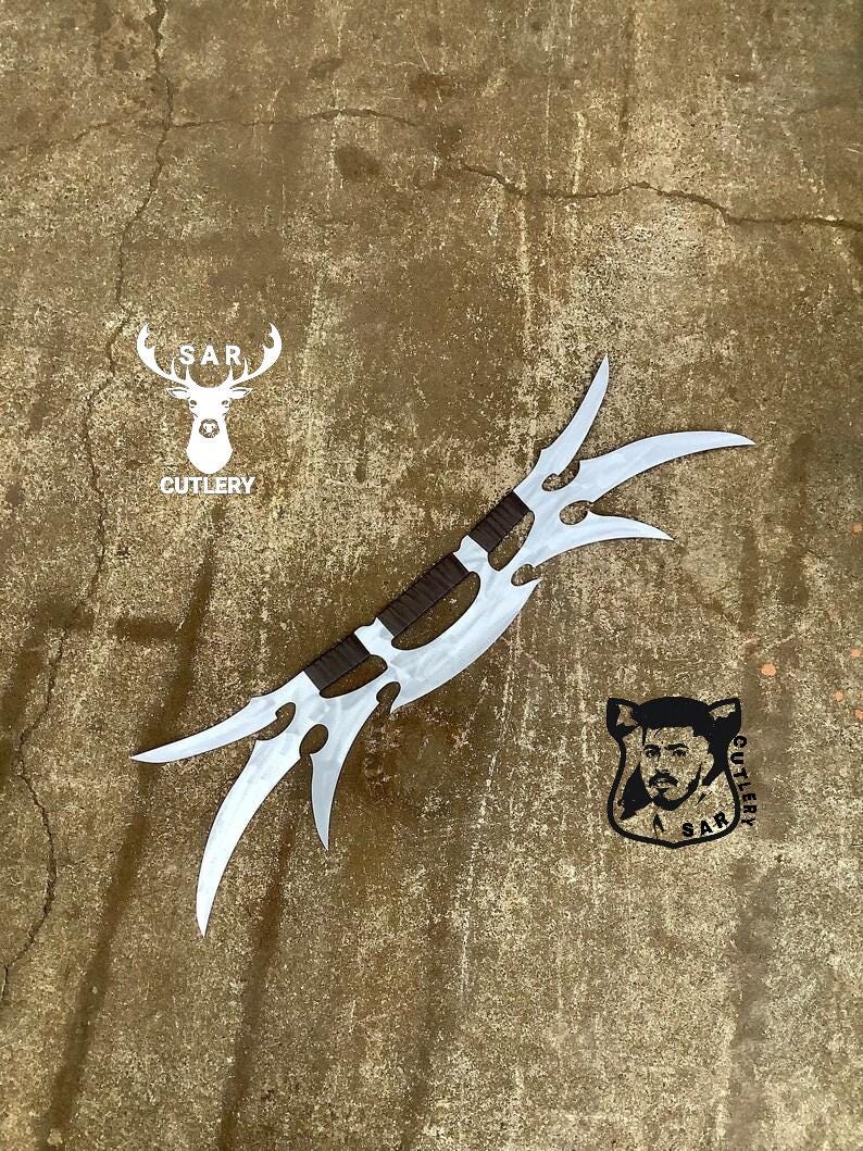May include: A silver-colored, double-edged blade with three curved points on each side. The handle is wrapped in brown leather. The image also includes the text "S.A.R. CUTLERY" and a logo of a deer.