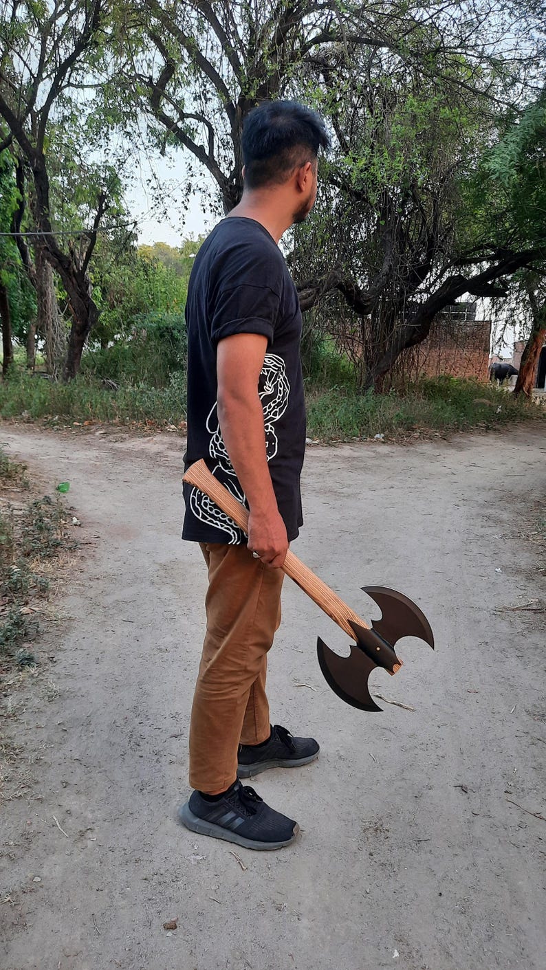Puede incluir: Un hacha de batalla de doble filo con un mango largo de madera clara con veta visible y dos cabezas de hacha sim&eacute;tricas de metal negro mate. El hacha se sostiene a un lado, mostrando sus hojas curvas y los remaches met&aacute;licos centrales.