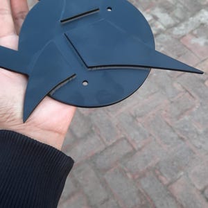 May include: A black, geometric metal sculpture with a circular base and angular, pointed extensions. The design features sharp lines and a symmetrical arrangement, held in a hand against a brick background.
